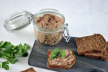 On a wooden board, several slices of black bread with liver pate. Next to a glass jar with pate. In the frame, green parsley. Light background.  Close-up. Macro photography.