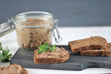 On a wooden board, several slices of black bread with liver pate. Next to a glass jar with pate. In the frame, parsley greens and a knife. Light marble surface. Close-up. Macro photography.