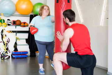 Fototapeta na wymiar Full length portrait of obese young woman hitting punching bag in gym during personal training with fitness instructor