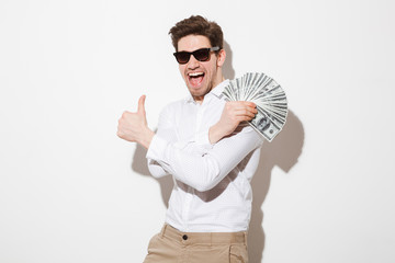 Joyous man in shirt and sunglasses rejoicing while demonstrating fan of money in dollar currency and showing thumb up, isolated over white wall with shadow