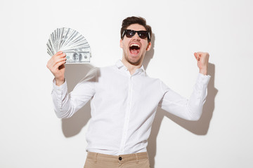 Emotional brunette guy in shirt and sunglasses shouting with clenched fist while holding money prize in cash, isolated over white wall with shadow