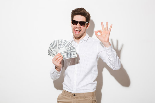 Photo of happy winner man in shirt and sunglasses smiling holding fan of money in dollar banknotes and showing ok symbol, isolated over white wall with shadow