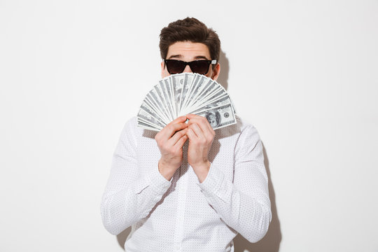 Photo of funny man in casual shirt and sunglasses covering his face with fan of money in dollar currency, isolated over white wall with shadow