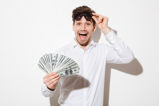 Splendid photo of excited man in shirt taking off black sunglasses and enjoying fan of money dollar bills with pleasure and joy, isolated over white wall with shadow