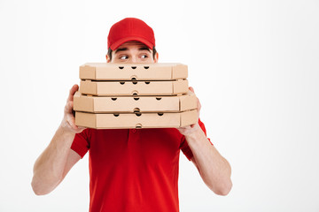 Image of joyful delivery man in red uniform covering face with stack of pizza boxes and looking aside, isolated over white background