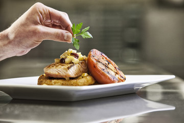 Close-up of a chef's hands adding a garnish to a salmon dinner plate.