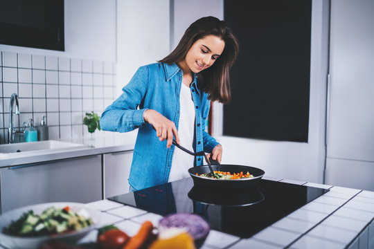 Beautiful young brunette woman cooking vegetarian food in the kitchen at home, happy smiling girl preparing organic vegetables in frying pan, healthy food lifestyle