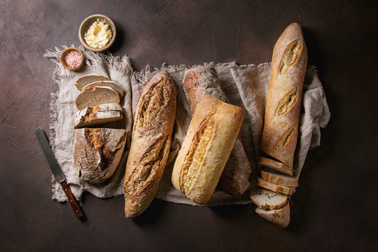 Variety of loafs fresh baked artisan rye, white and whole grain bread on linen cloth with butter, pink salt and vintage knife over dark brown texture background. Top view, copy space.