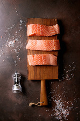 Sliced raw uncooked salmon filleton wooden cutting board with sea salt and pepper over dark brown texture background. Top view, space.