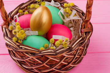 Easter eggs and pussy-willow in woven basket. Gold egg and colorful eggs decorated with pussy-willow twig in basket on pink wooden background.