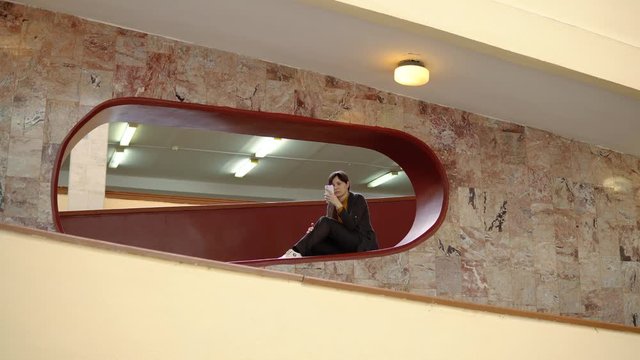 A young attractive woman sits in a niche and prints a message using a smartphone indoors with marble walls.