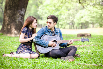 Fototapeta premium Asian couple in love playing acoustic song guitar sitting on grass in the park