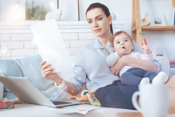 Family and career. Good looking nice confident woman holding a document and reading it while sitting with her child on the sofa