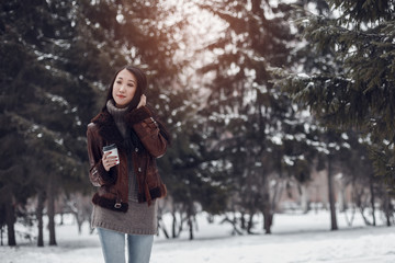 Fototapeta na wymiar Portrait of young beautiful smiling Asian girl woman in brown knitted sweater and boho leather coat walking outdoors in winter fir park holding a take away cup of coffee or tea
