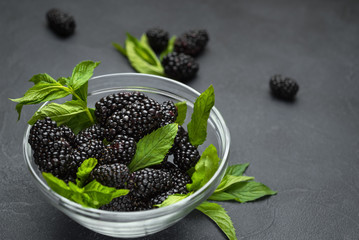Glass plate with ripe blackberries and green mint leaves on a black surface.