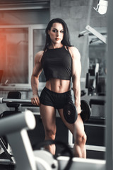 Naklejka na ściany i meble Full length portrait of a young attractive sexy beautiful fitness girl in black tank top and shorts standing in gym near training apparatus showing her press and looking at camera. Daria Manyilenkova