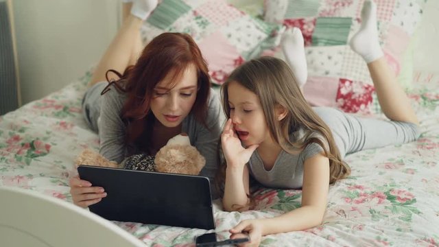 Beautiful young mother and her cute daughter in pajamas laughing and looking in digital tablet while lying on bed at home in the morning