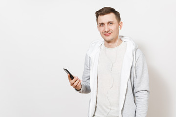 Young handsome smiling man in t-shirt, light sweatshirt listening to music on earphones on mobile phone isolated on white background. Concept of emotions, good mood. Copy space for advertisement.