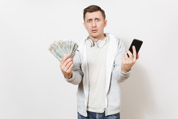 Young handsome man in t-shirt and light sweatshirt with headphones around neck holds bundle of dollars, cash money, mobile phone in hands in perplexity isolated on white background. Concept of success