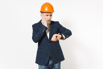 Irritated dissatisfied businessman in dark suit, protective hardhat looking on clock on hand and clinging to head isolated on white background. Time is running out. Male worker for advertisement.