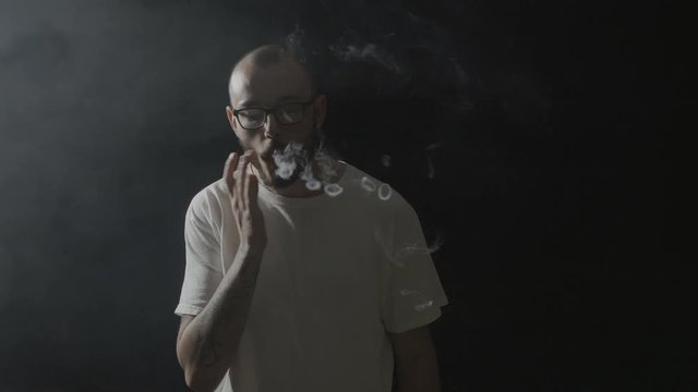 Young man wearing glasses inhaling smoke and practicing various vaping tricks making circles in a dark room