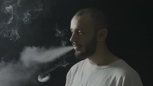 Portrait of an attractive teenager boy vaping and making smoke tricks on a dark background
