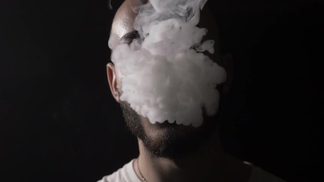 Bold young male musician vaper with beard vapes and blows on his mouth a huge cloud of vape smoke that covers his face in slow motion