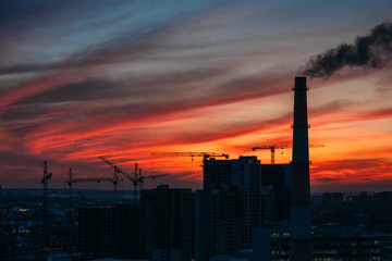 Fototapeta na wymiar Panorama of city sunset and silhouettes of cranes, high-rise buildings and construction site with smoke
