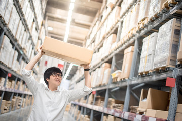 Young Asian man carry paper box over head between row of shelves in warehouse, shopping warehousing or working pick and packing concepts