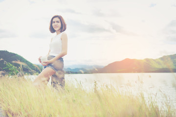 Beautiful woman in white cloth sitting and holding a hot coffee in white cup in field at giant lake with her vacation time.