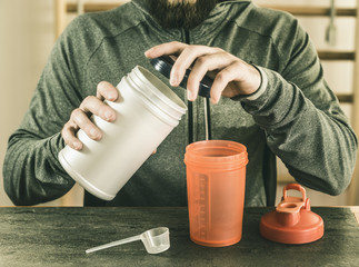 young man with a beard, in sports clothes, opens a can of protein, against the background of sports equipment