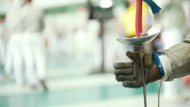 Young fencer turns a rapier in his hand, in a gym