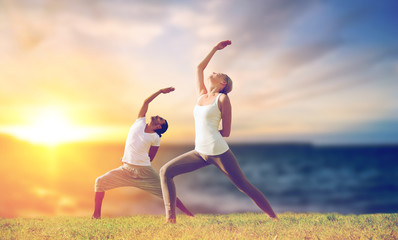 Fototapeta na wymiar fitness and people concept - couple making yoga warrior pose outdoors over sea background