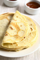 Freshly made crepes stacked on plate with strawberry and apricot jam in the back, photographed with natural light (Selective Focus, Focus in the middle of the image)