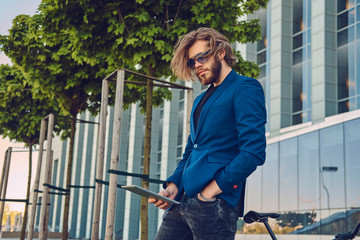 A handsome fashionable bearded male with long hair dressed in stylish clothes, working with a tablet computer after bicycle riding.