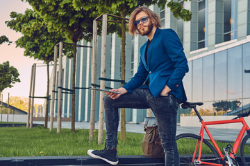 A handsome fashionable bearded male with long hair dressed in stylish clothes, working with a tablet computer after bicycle riding.