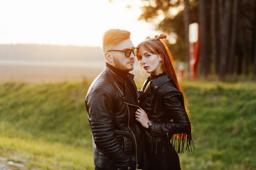 Fototapeta premium slim stylish girl with long red hair hugging a brutal guy with a beard and wearing sunglasses at sunset