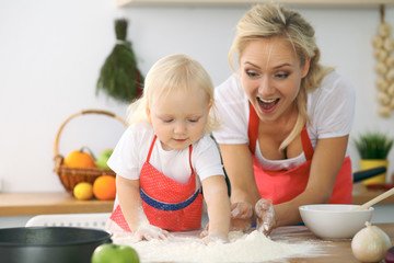Little girl and her blonde mom in red aprons  playing and laughing while kneading the dough in the kitchen. Homemade pastry for bread, pizza or bake cookies