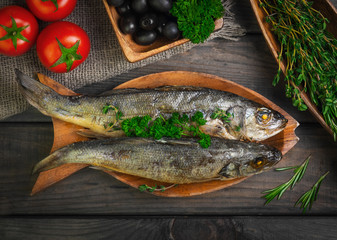 Two grilled whole fish trout on gray wooden background. Additional ingredients for grilled fish greens rosemary, thyme, parsley, tomatoes, olives. Top view.