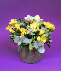 Festive flower arrangement of yellow and white freesia flowers and other plants with Easter eggs decorated on violet background.