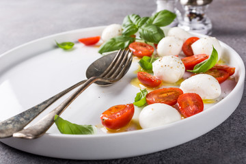 White plate of healthy classic delicious caprese salad with ripe tomatoes and mozzarella cheese with fresh basil leaves on gray concrete background with space for text. Italian food.