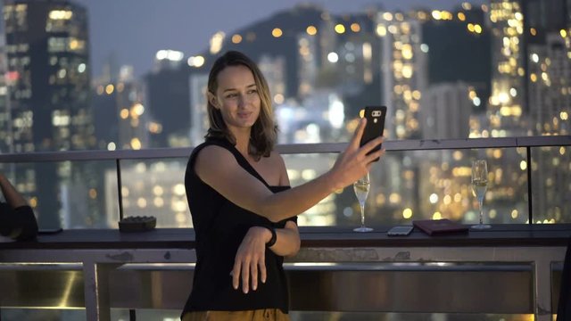 Happy woman drinking champagne and taking selfie photo with cellphone in skybar, luxury bar at night
