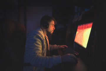 A cute male gamer sits in a cozy room behind a computer and plays games. Gamer plays horror game at night. Computer games. Gamer concept. © bodnarphoto