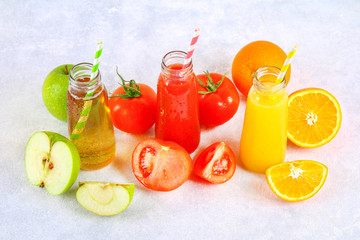 Bottles with fresh orange, apple, tomato juice and colored tubules on a gray concrete table.