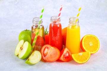 Bottles with fresh orange, apple, tomato juice and colored tubules on a gray concrete table.