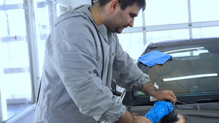 A man in a special suit polishes a gray car body, a tool for polishing cars, into a workshop.