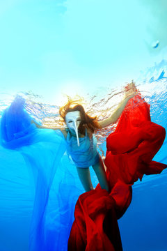 Girl in a white mask with a long nose swims underwater and plays with red and blue fabrics. Portrait. Vertical orientation