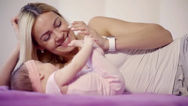 blonde woman is playing with her infant daughter lying on a bed in room, kissing her little feet