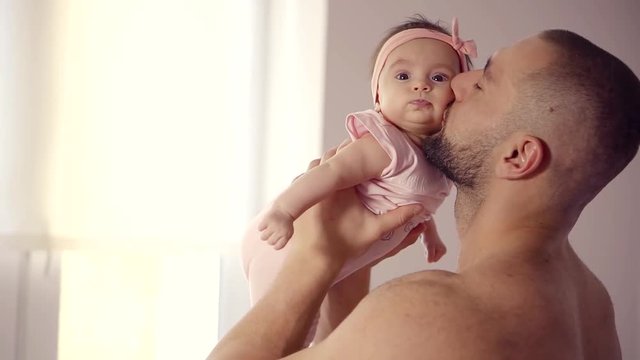 adult man is playing with his baby, holding daughter in hands, kissing in cheek and speaking with her funny