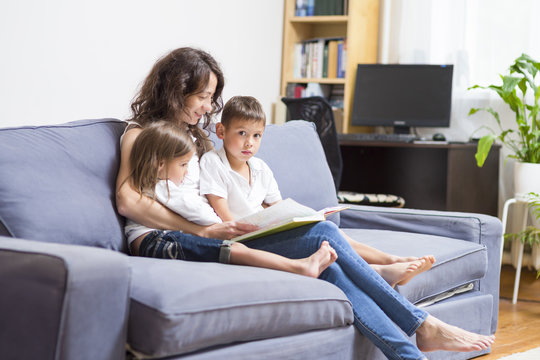 Family Relationships Concepts. Mother With Her Two Children Reading A Book Together. Sitting On Couch Indoors.